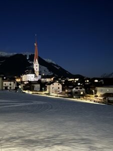 Verschneites Dorf in den Alpen bei Nacht mit einer beleuchteten Kirche und tiefblauem Himmel.